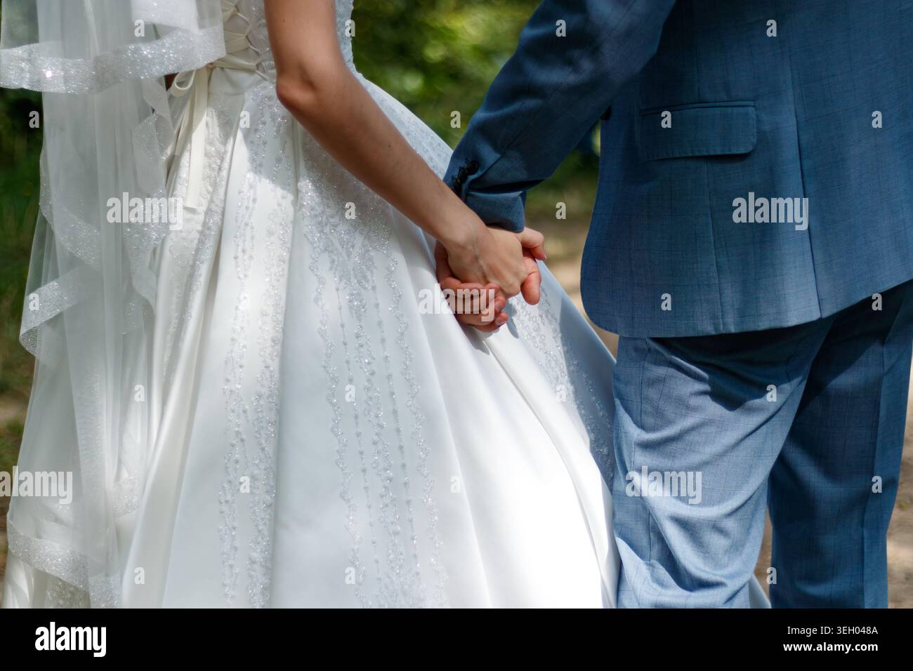 A couple walks hand in hand on a path surrounded by trees. They are dressed in formal wedding attire. Sunlight filters through the leaves above. The s Stock Photo