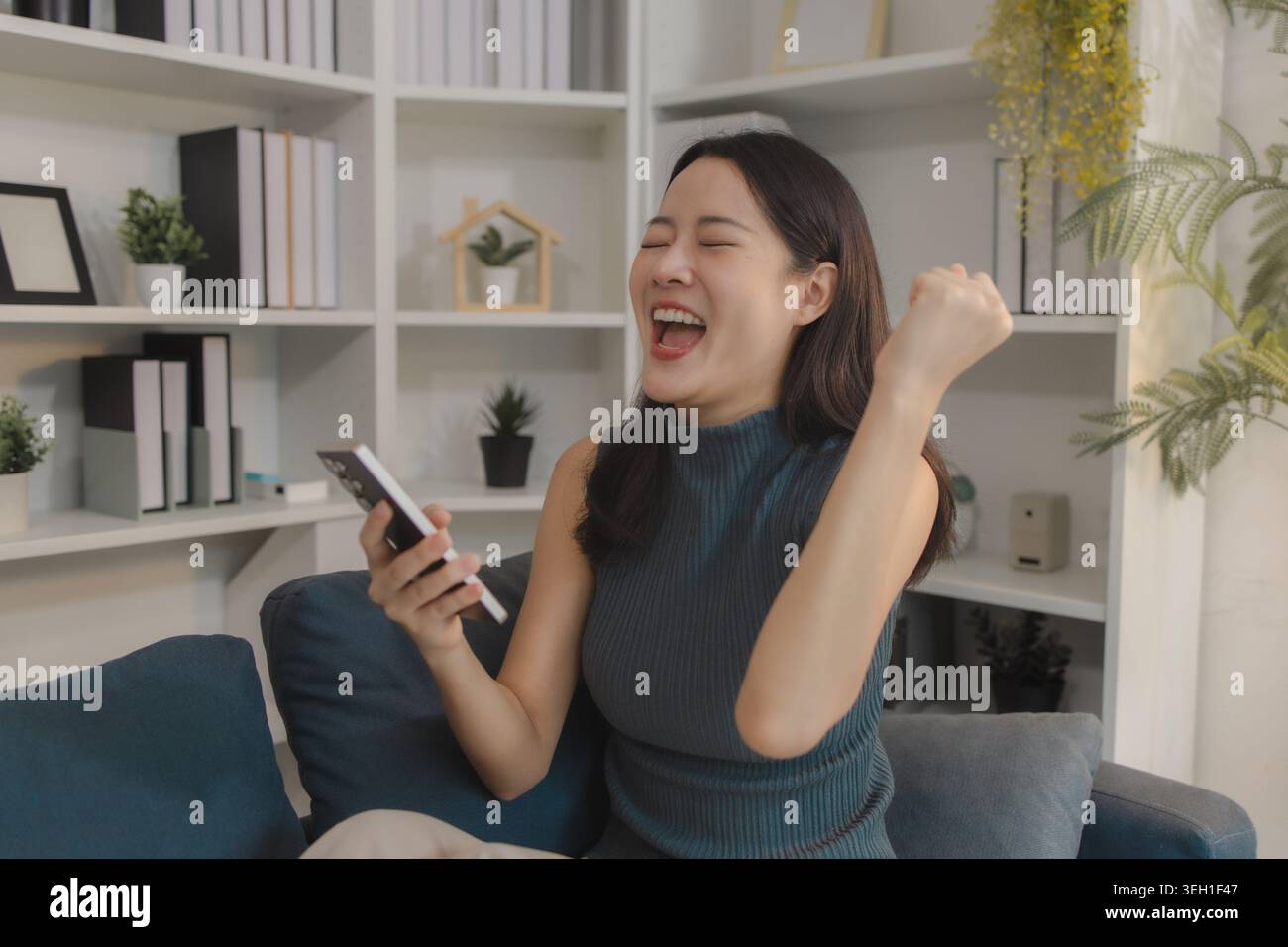 Young Asian woman sitting on sofa celebrating while using smartphone, expressing excitement and success, representing positive online experience, digi Stock Photo