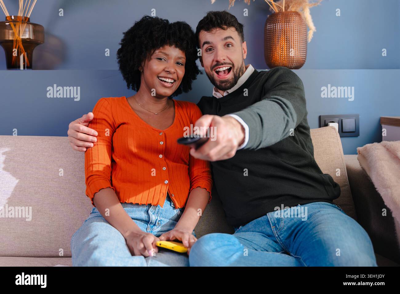Happy multiracial couple watching television and using remote control on sofa Stock Photo