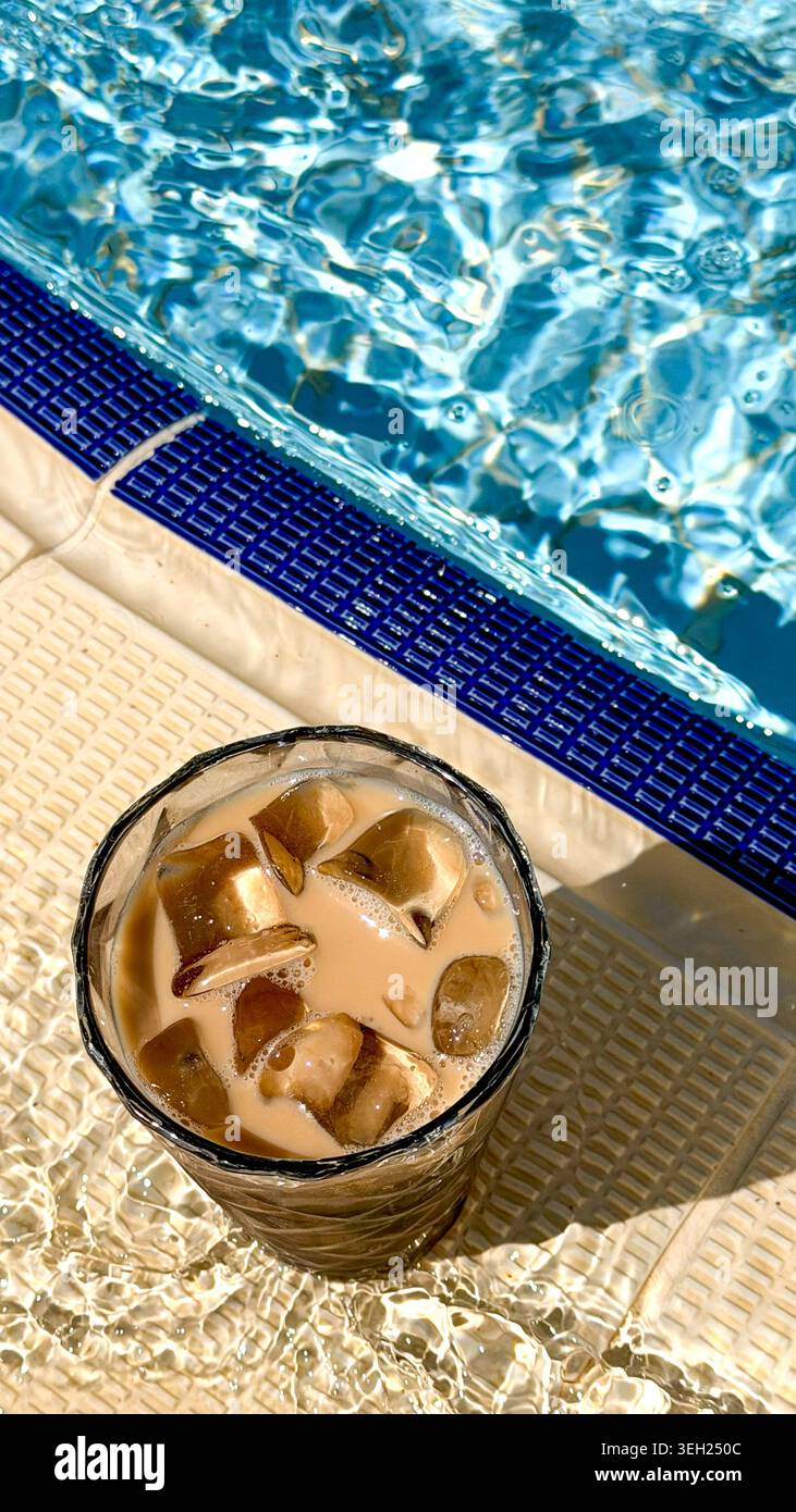 Iced coffee drink with ice cubes in a glass placed beside a swimming pool with clear blue water reflecting sunlight on a warm day Stock Photo