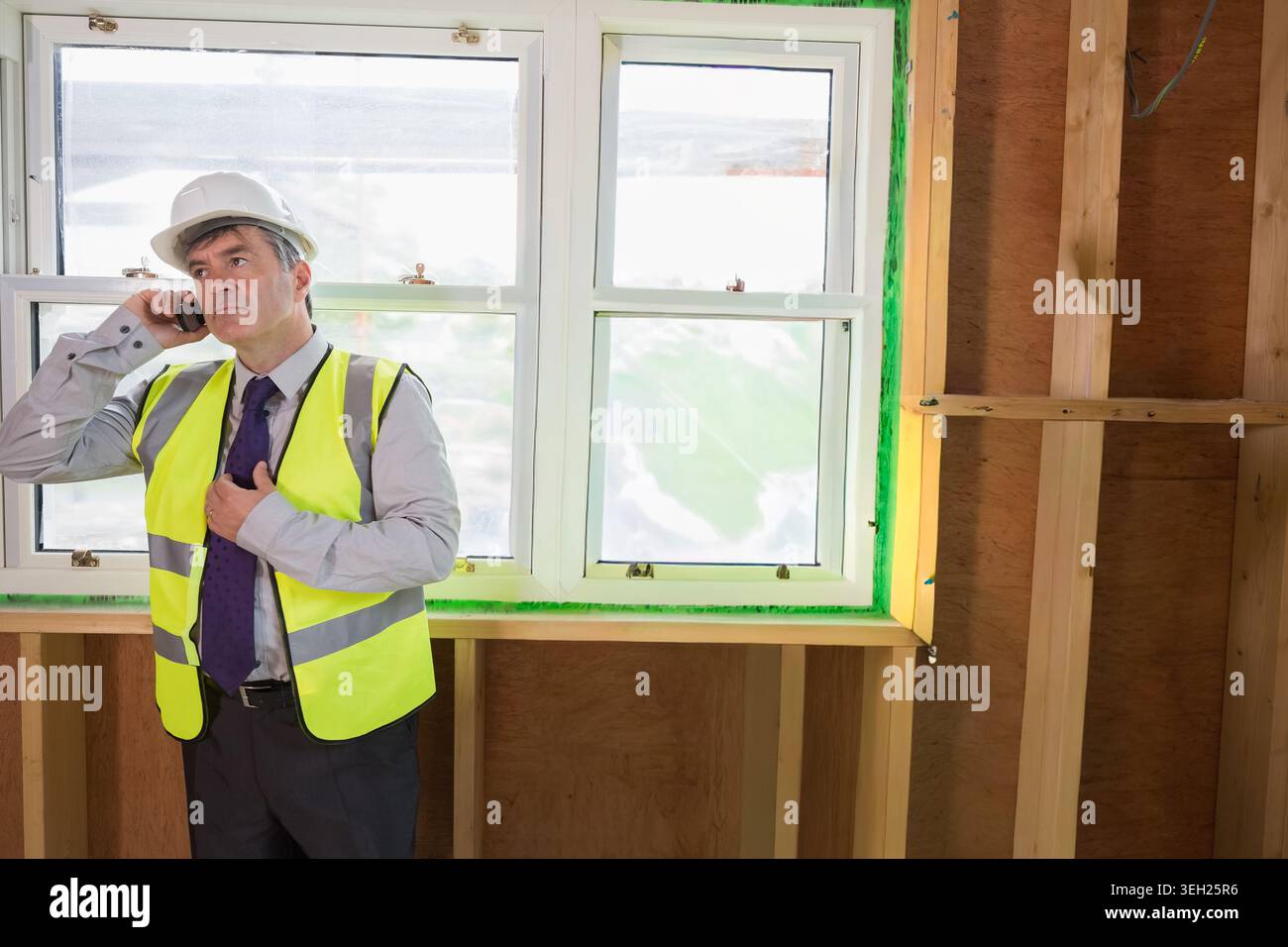 Mature man wearing hard hat, yellow vest, standing by sealed window holding phone and adjusting tie Stock Photo