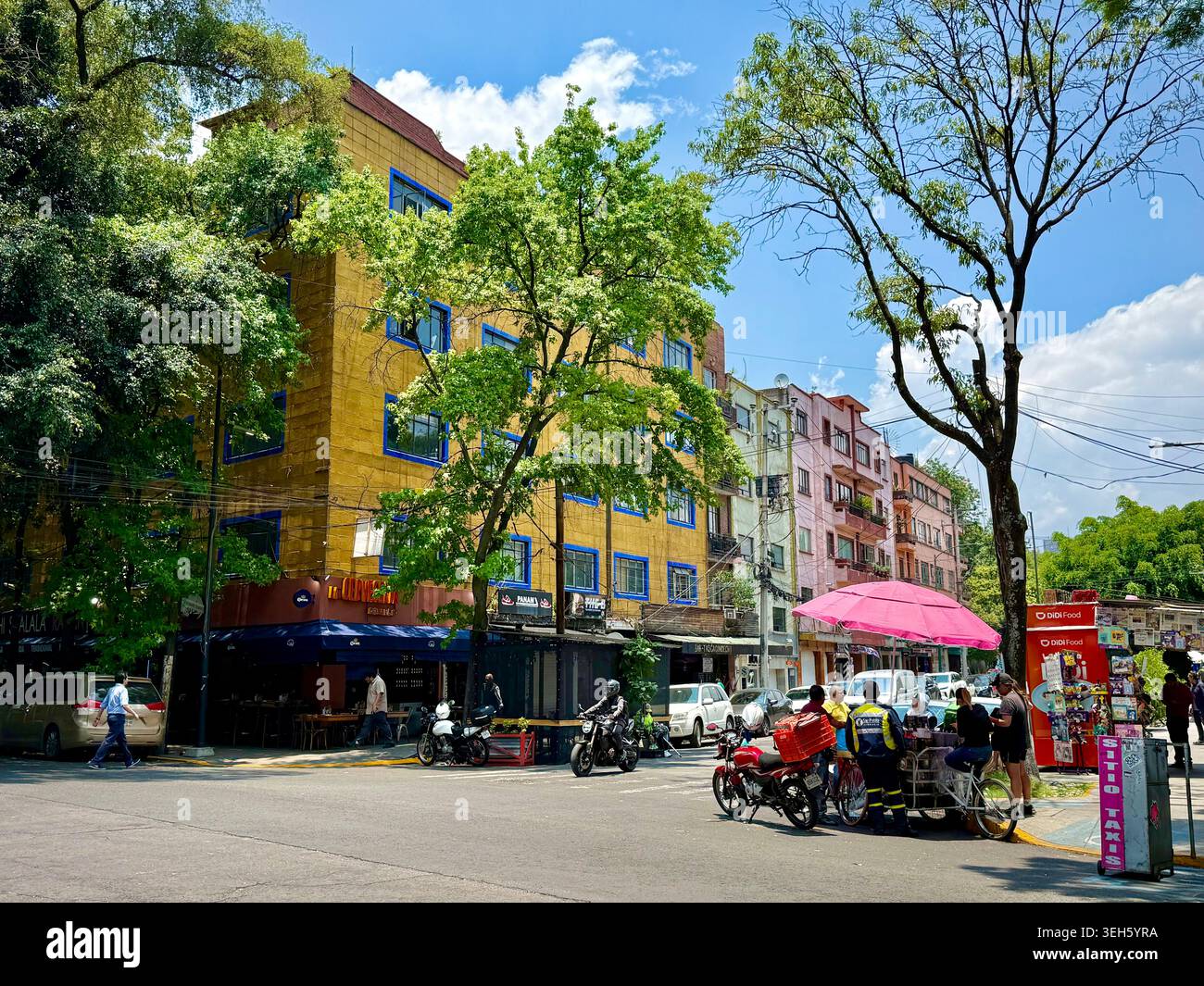 A lively street corner in La Condesa, Mexico City, with colorful apartment buildings, trees, motorcycles and a small street stand. Stock Photo