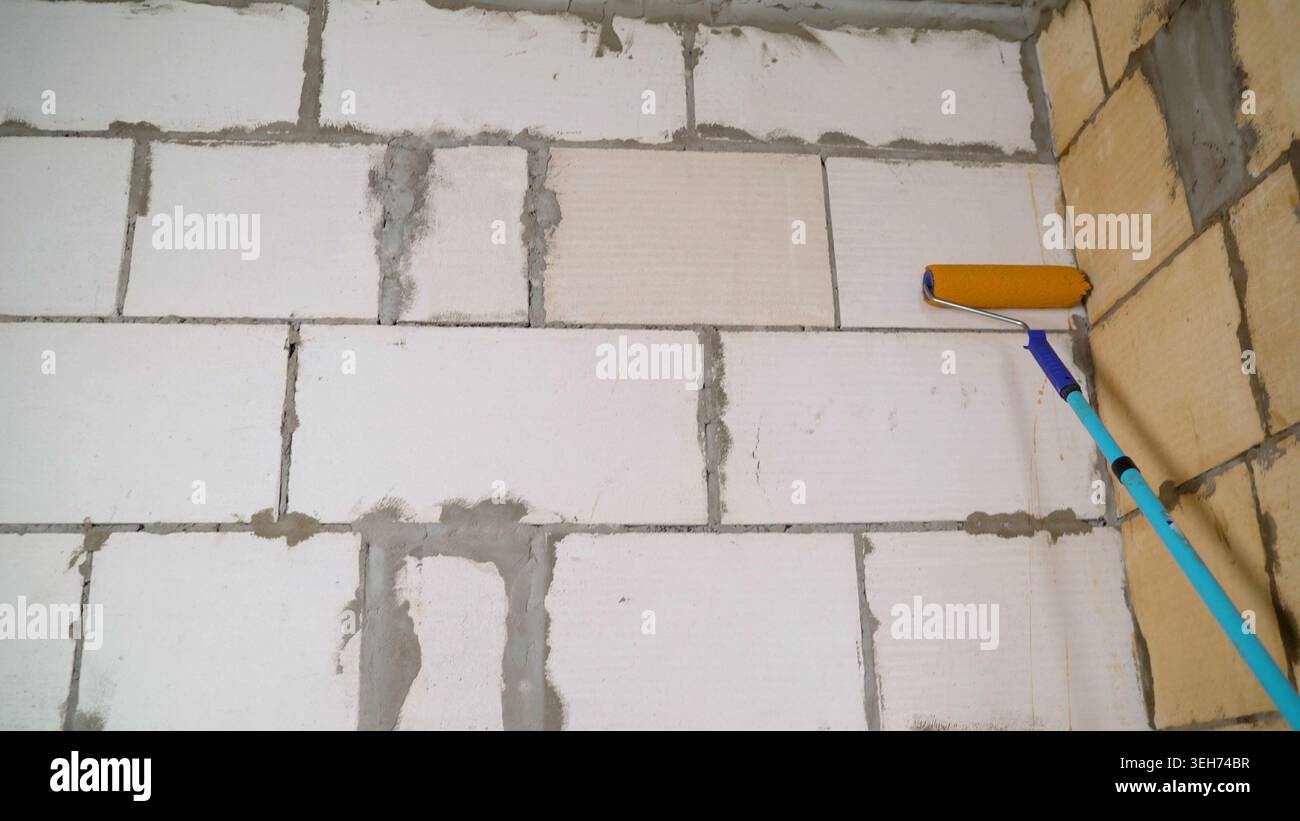 Close up of priming process on unfinished concrete block wall. Yellow roller applies beige coating. DIY home renovation and construction background. N Stock Photo
