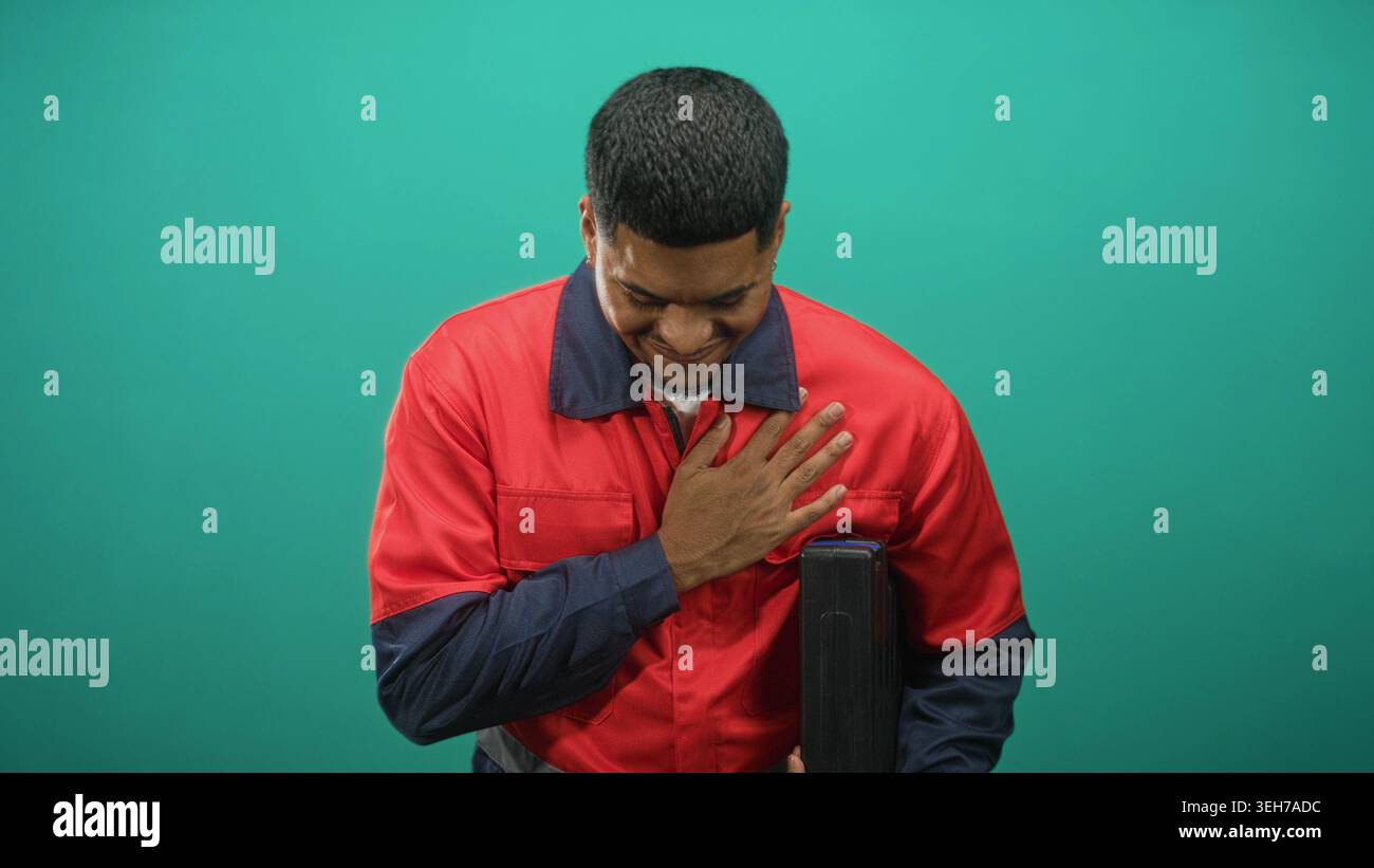 Hispanic bearded man in red work uniform stands holding a toolbox with one hand and places his hand on chest in a studio setting; pride duty. Stock Photo