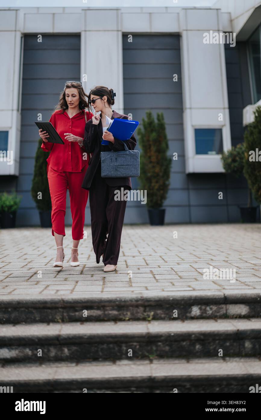 Two businesswomen walking and discussing documents outside a modern office building Stock Photo