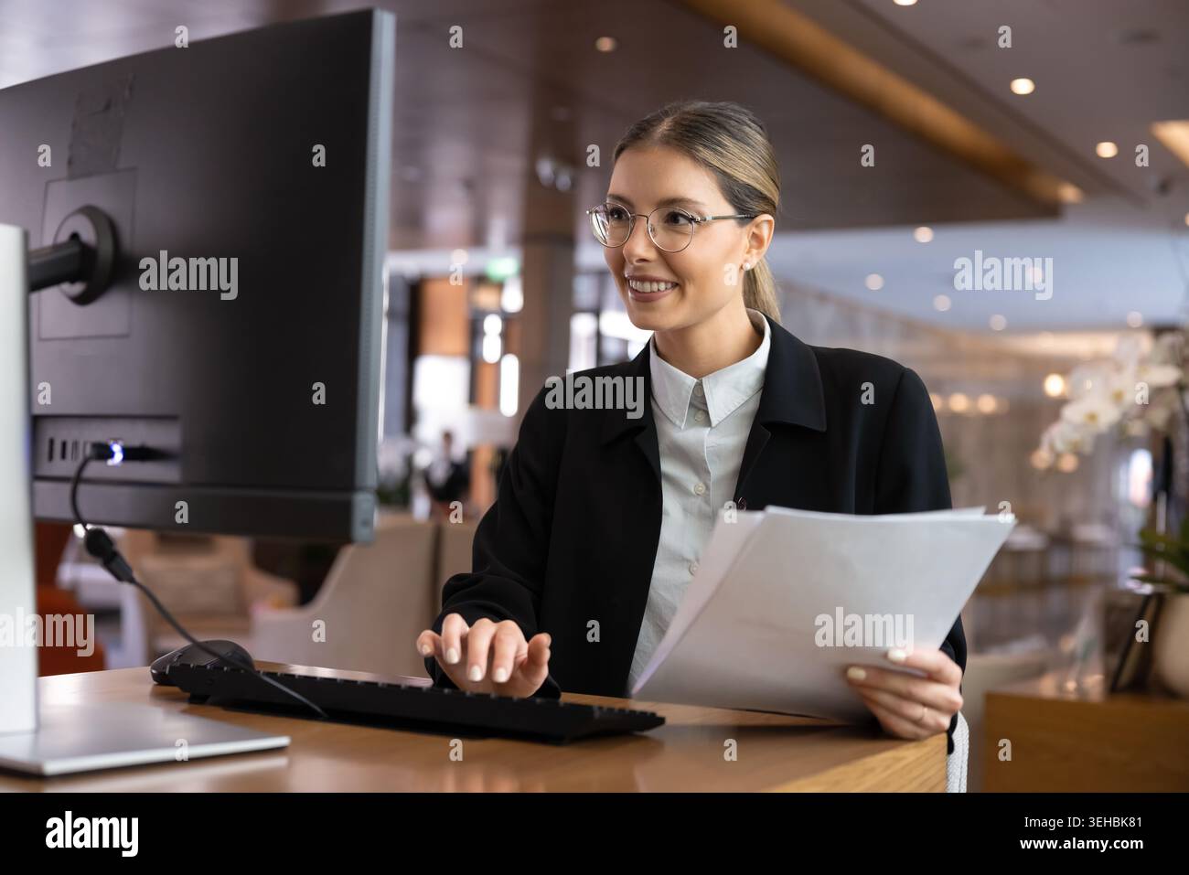Woman receptionist holding documents, working on computer at reception desk Stock Photo