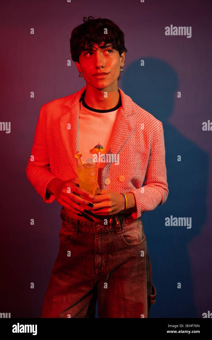 Young non-binary person holding a fun cocktail, celebrating personal style and identity in a vibrant setting Stock Photo