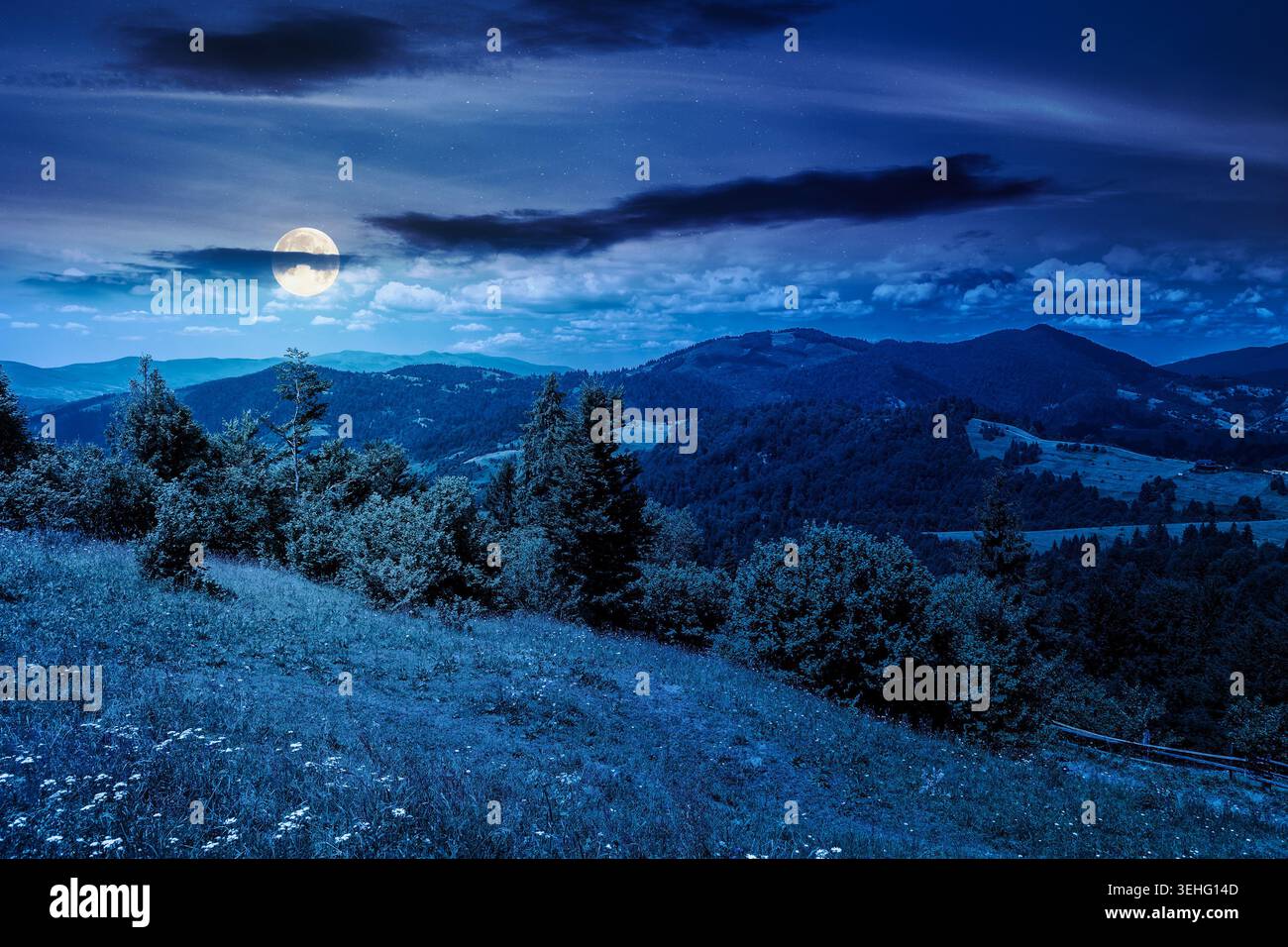 mysterious field on hillside in mountains near village at night. full moon over carpathian mountain landscape in summer. countryside view of alpine fi Stock Photo