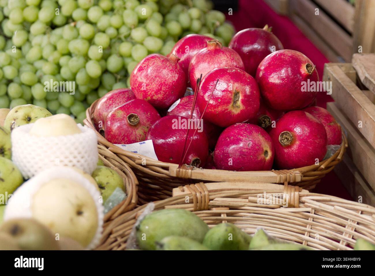 A wicker basket filled with bright red pomegranates displayed on a market table alongside green grapes and wrapped pears. Stock Photo