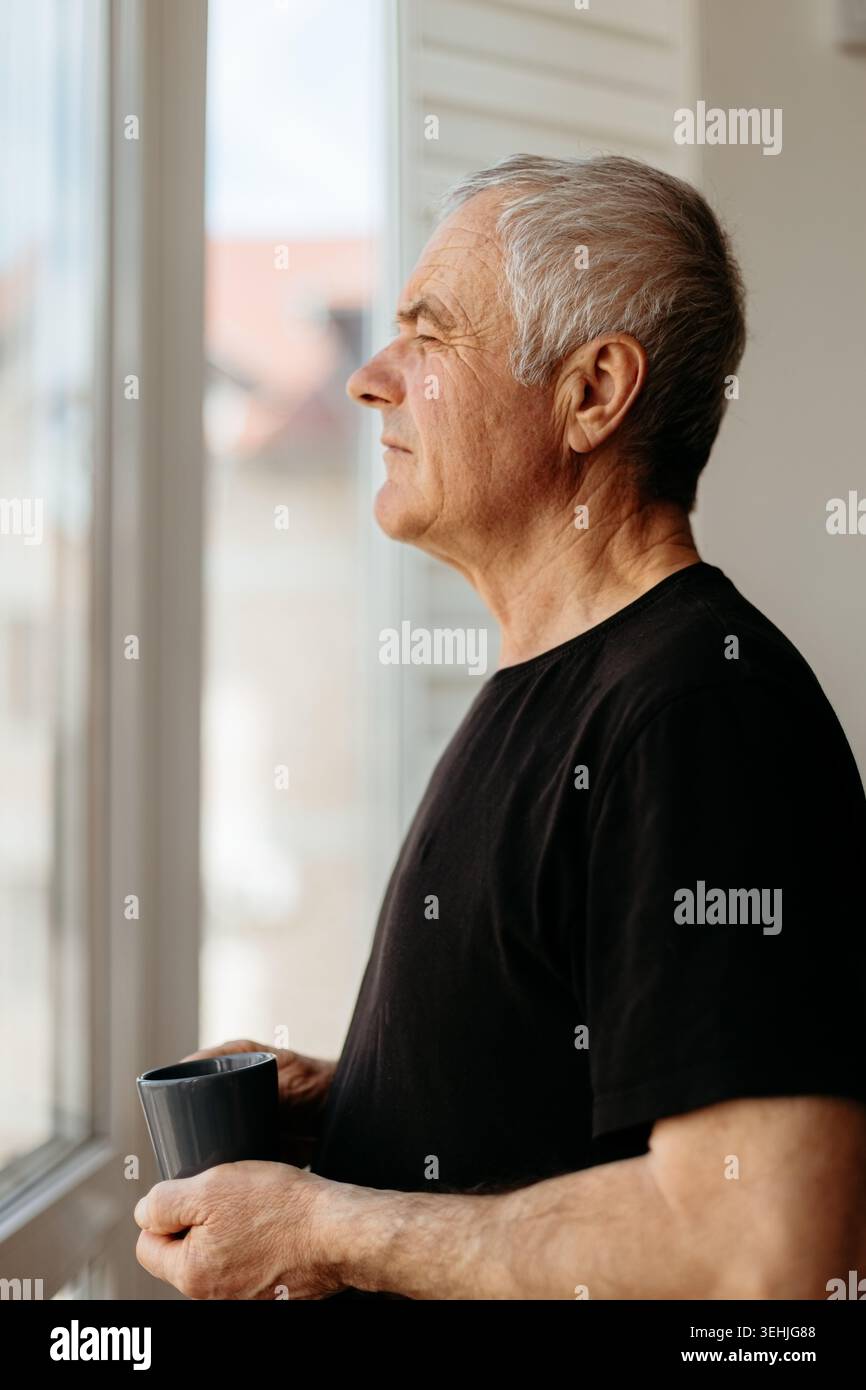 Senior man gazing thoughtfully through window while holding coffee cup Stock Photo