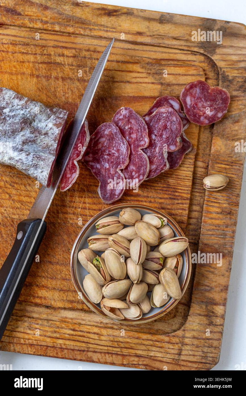 A knife, a piece of sliced salami, and a bowl of pistachios sit on a ...
