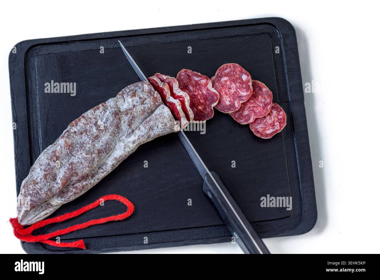 A whole salami and several slices lie on a black cutting board next to ...