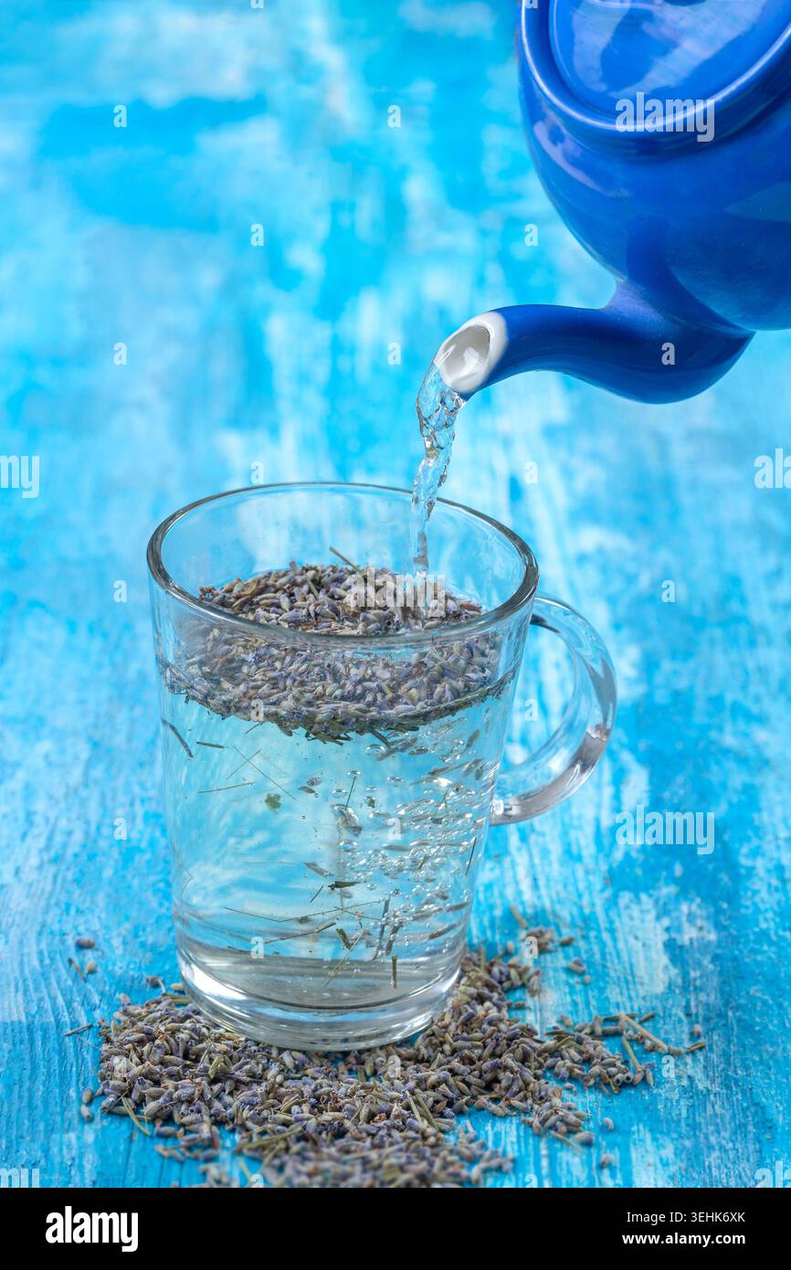 Water pours from a blue teapot into a glass mug containing dried purple ...
