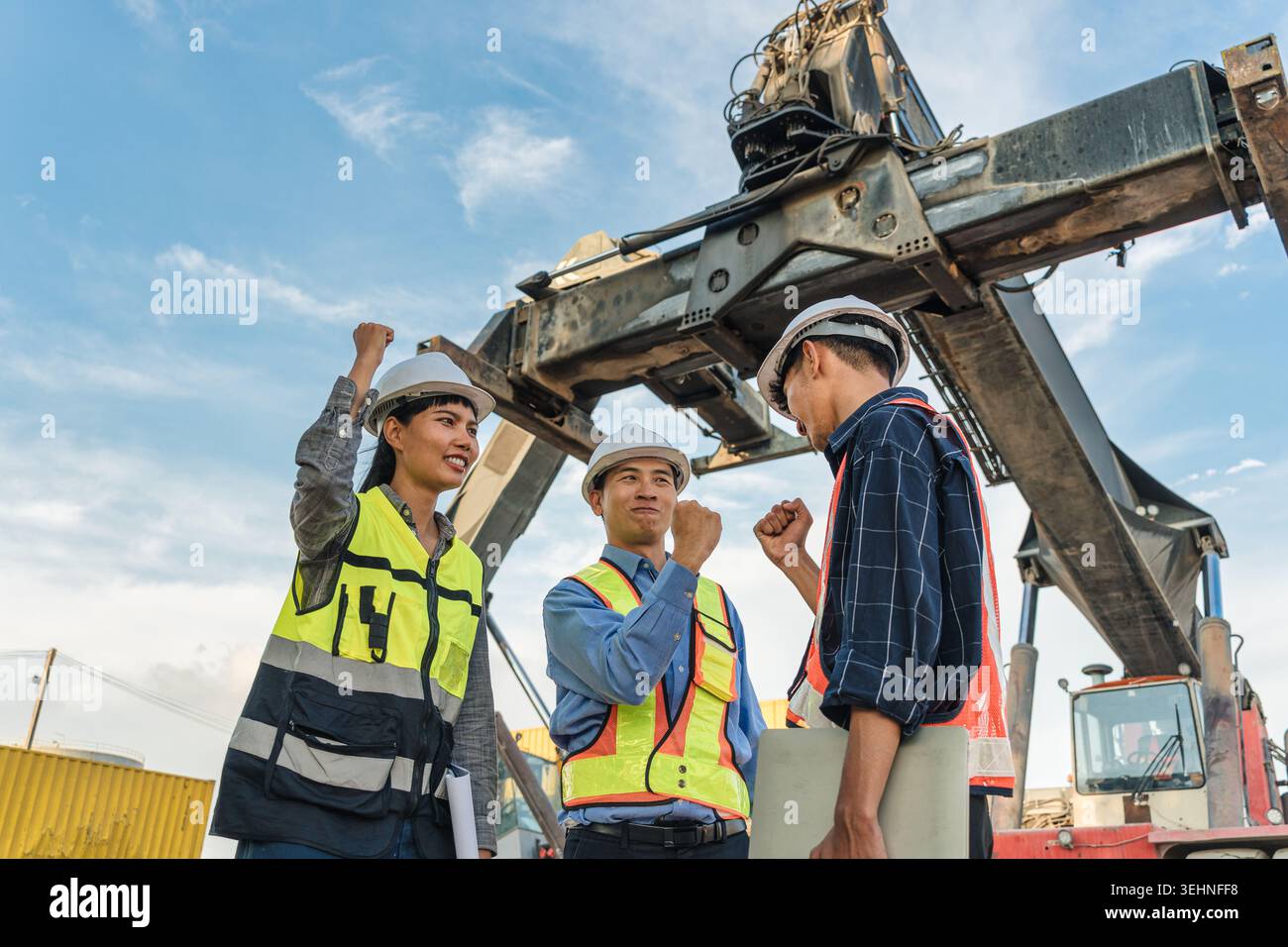 Three construction foreman workers wearing hard hat and safety vest celebrate with raised fist pump at industrial site with large crane machinery and Stock Photo