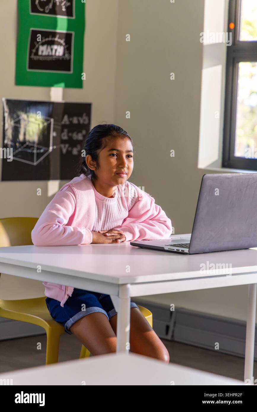 School-age Asian girl sitting on yellow chair in classroom, using silver laptop, wearing pink top Stock Photo