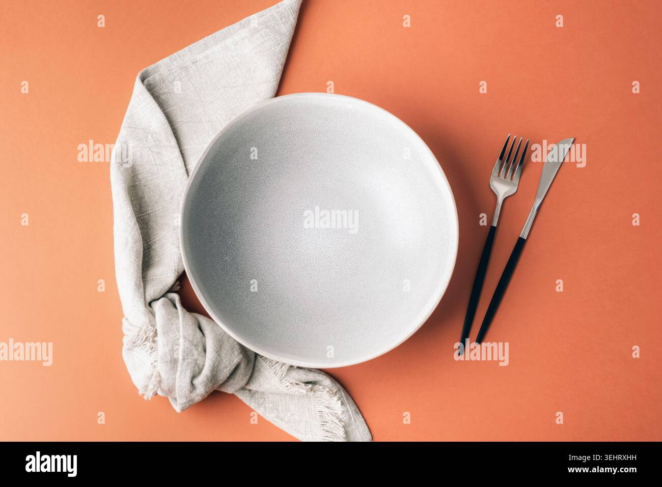 Minimal table setting with empty ceramic plate, linen napkin and modern cutlery on warm terracotta background. Elegant dining composition with copy sp Stock Photo