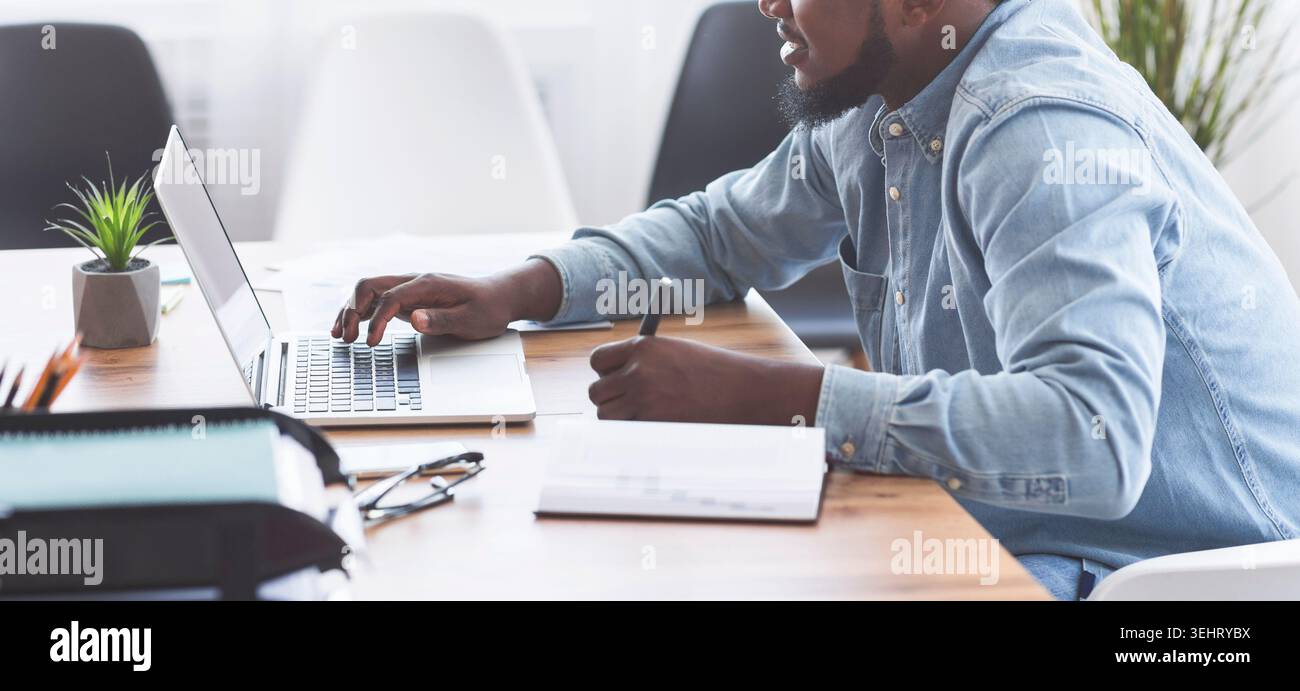 Black trainee taking notes while using laptop in office Stock Photo