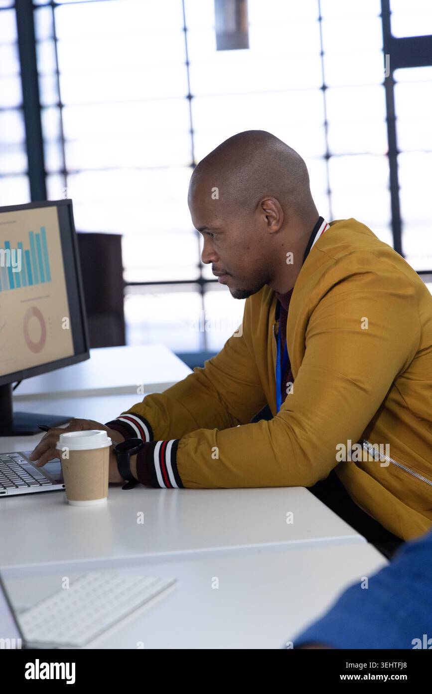 African American man wearing mustard jacket typing on laptop at desk with charts and coffee cup Stock Photo