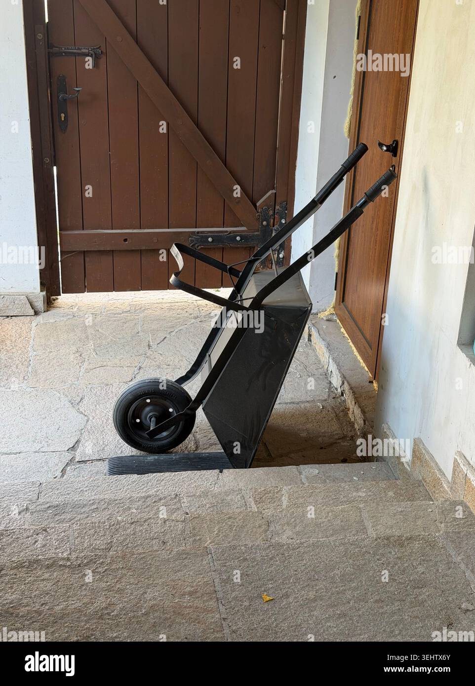 A black metal wheel barrow positioned leaning against a white wall on a stone covered pathway in a Bulgarian house Stock Photo