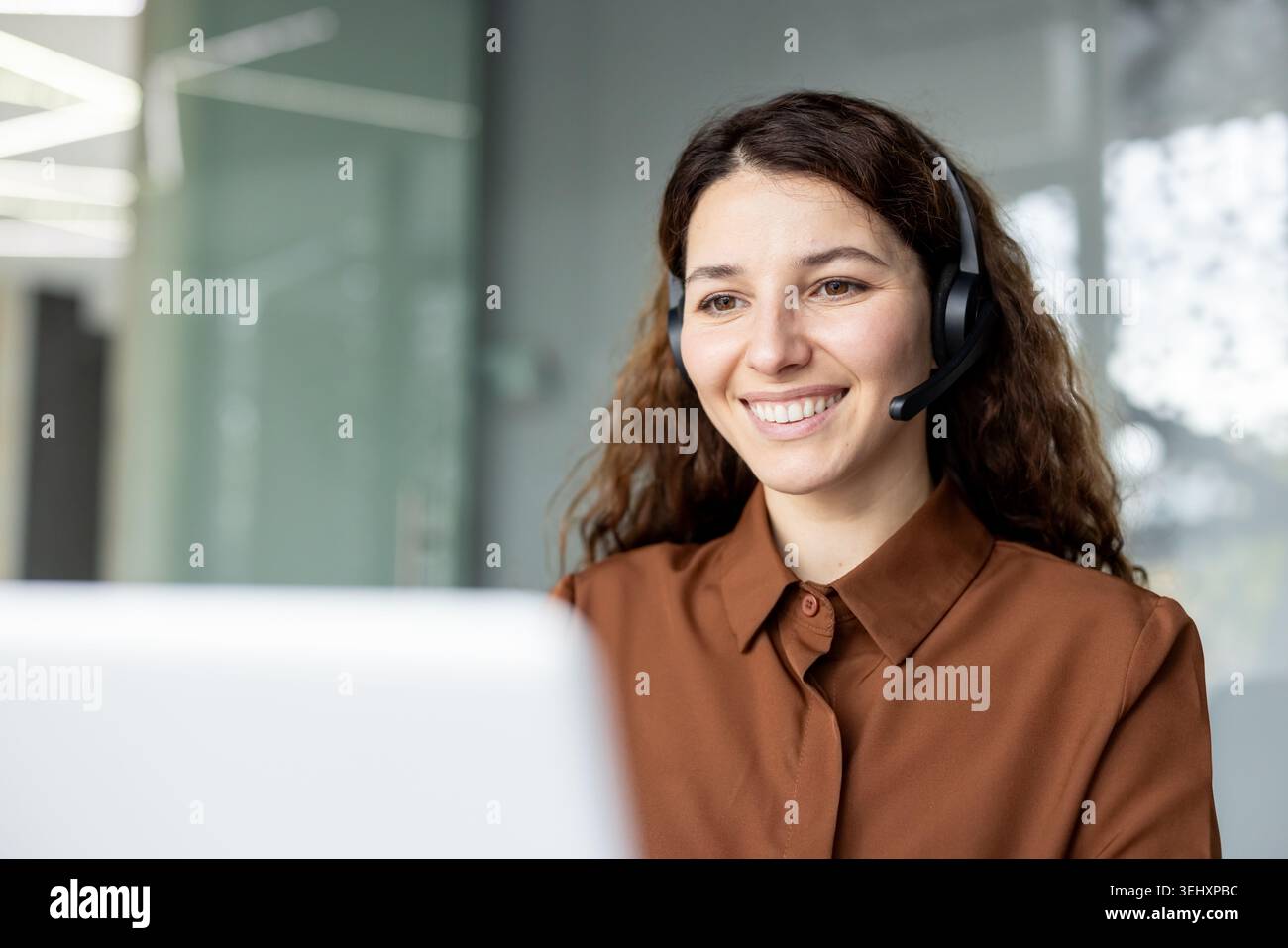 Young female call center agent wearing a headset and smiling, happy to help customers while working in a modern office environment or teleworking from home Stock Photo