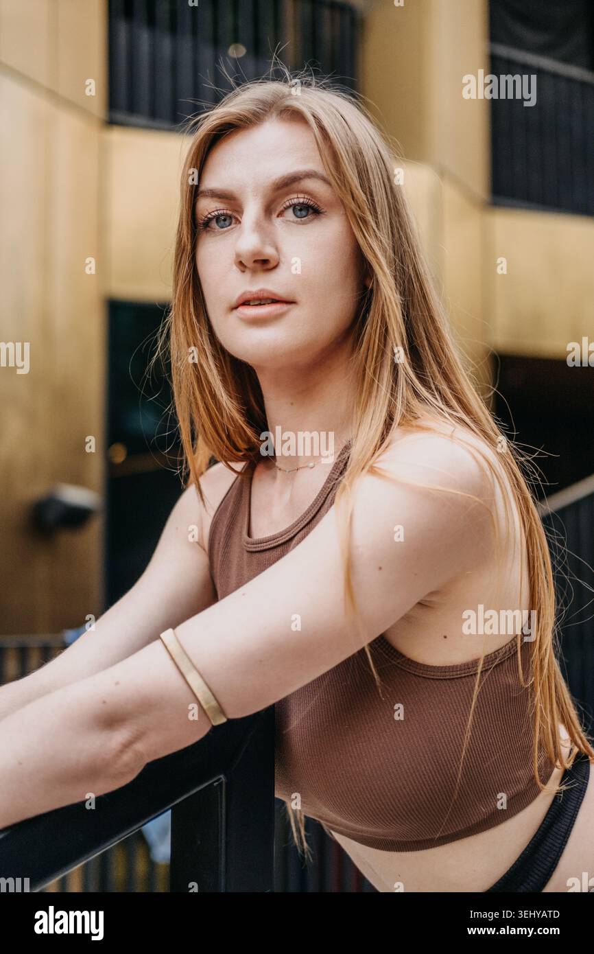 Woman leans on railing outside modern building looking directly at camera. Empowered women concept, self assured expression, authentic portraiture. Stock Photo