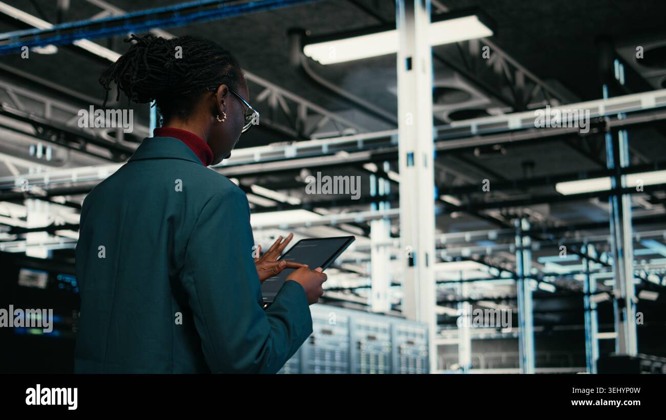 Data center programmer writing code for resource optimization using tablet application. African american woman walking in server hub using programming script on device to upgrade equipment, camera A Stock Photo