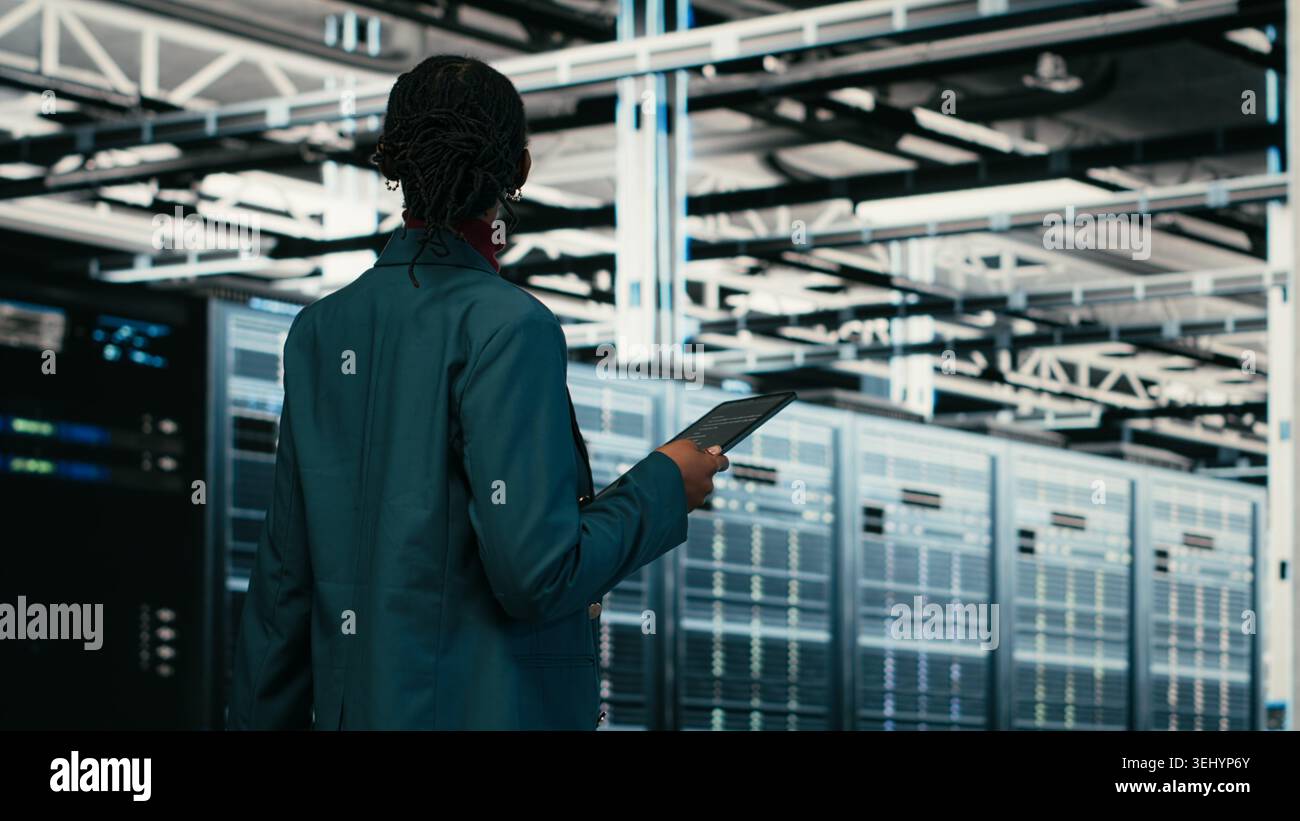 Female programmer walking in data center using tablet to write code, compiling software patches. African american woman in server hub using device application to run programming script, camera B Stock Photo