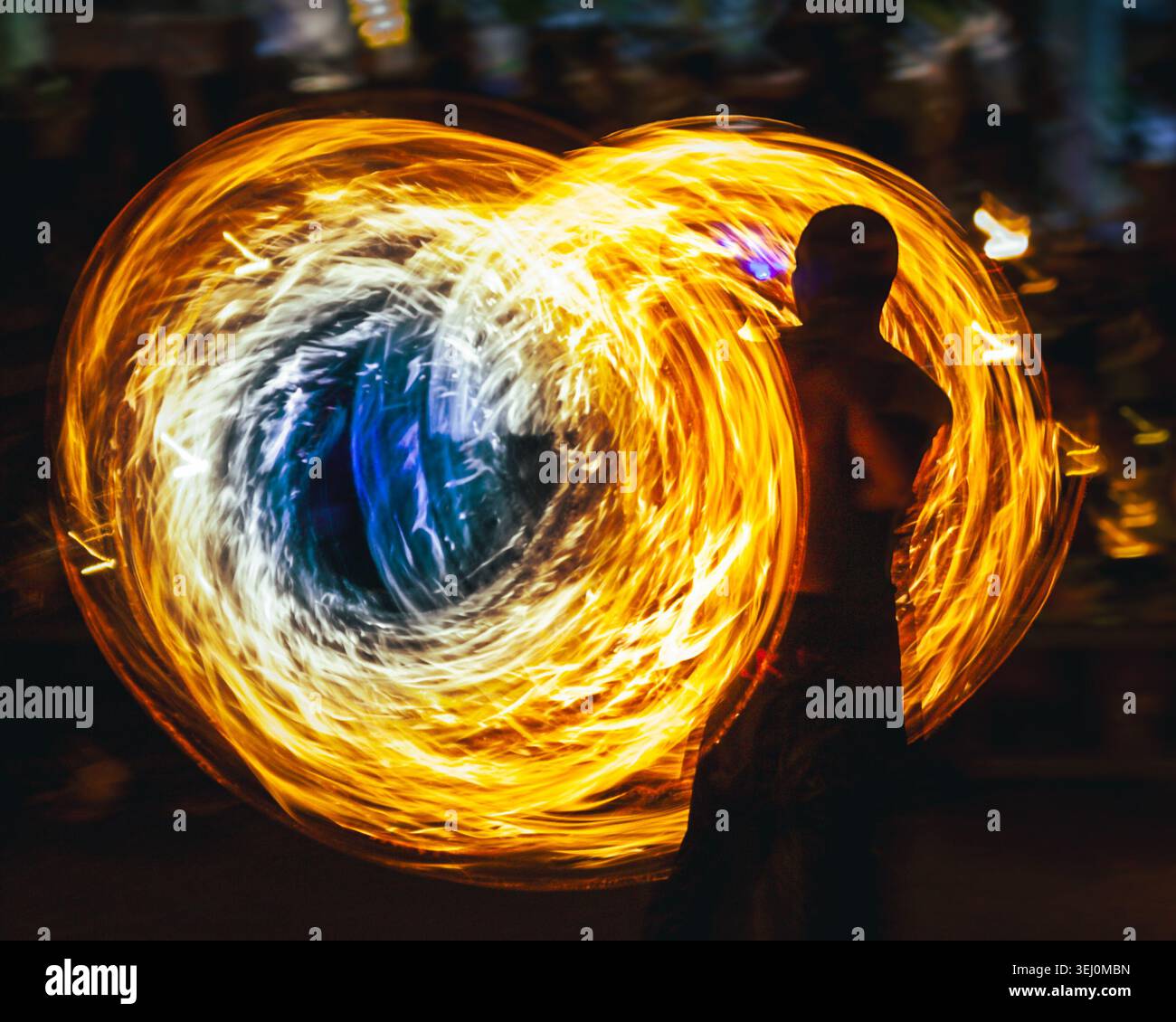 Person performing fire spinning with poi at night, creating abstract swirling light trails in vibrant orange, yellow, white, and blue hues, highlighting motion and energy Stock Photo