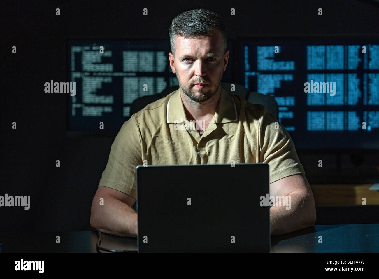 Hacker works in dark room alone. Hacker analyzes green code on laptop. Hacker monitors data flow in system. Hacker attempts network security breach. Stock Photo