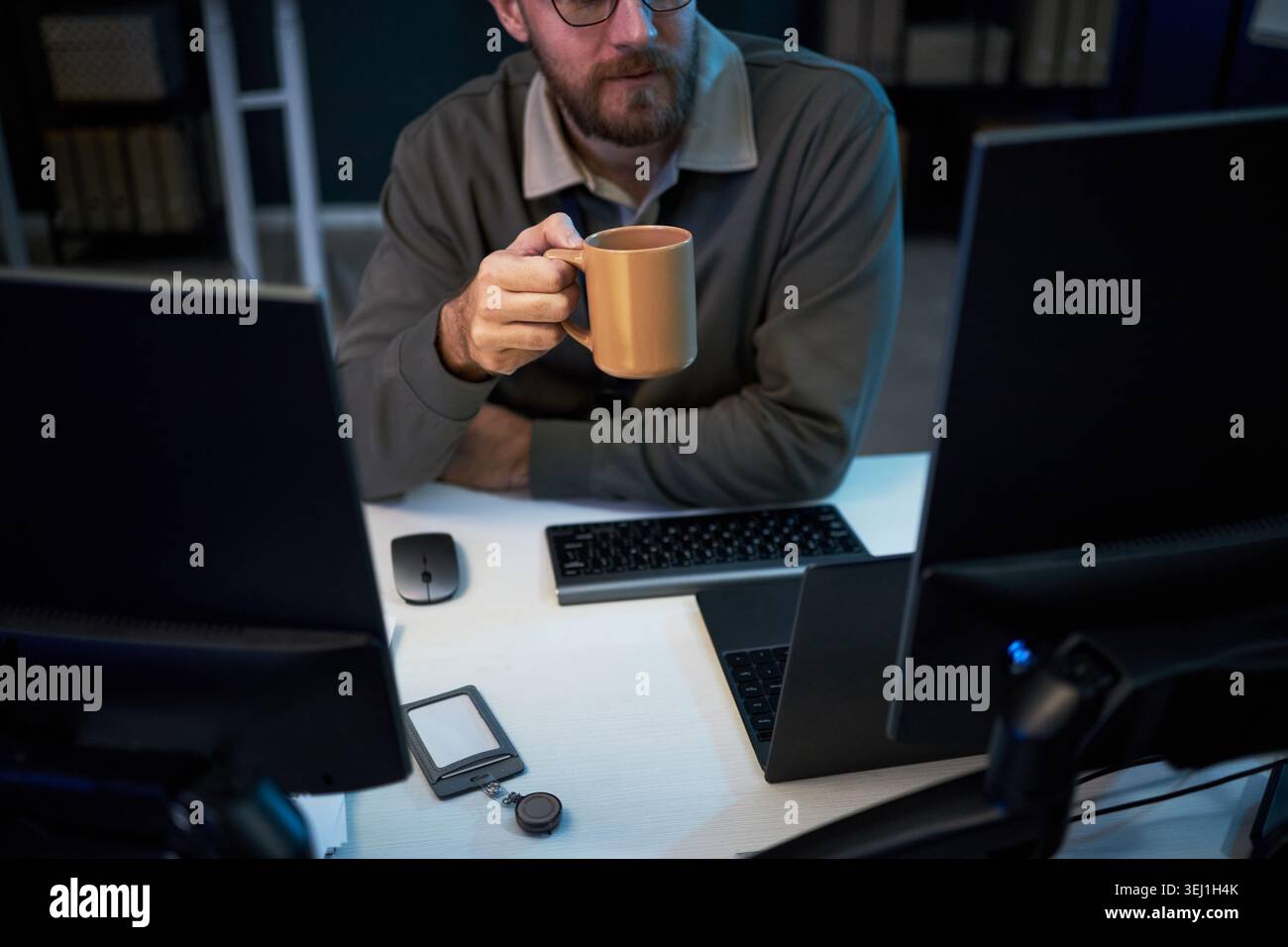 Middle aged Caucasian man holding coffee mug while working between multiple monitors and laptop at desk, suggesting late shift productivity. Ideal for remote work or tech workflow marketing Stock Photo