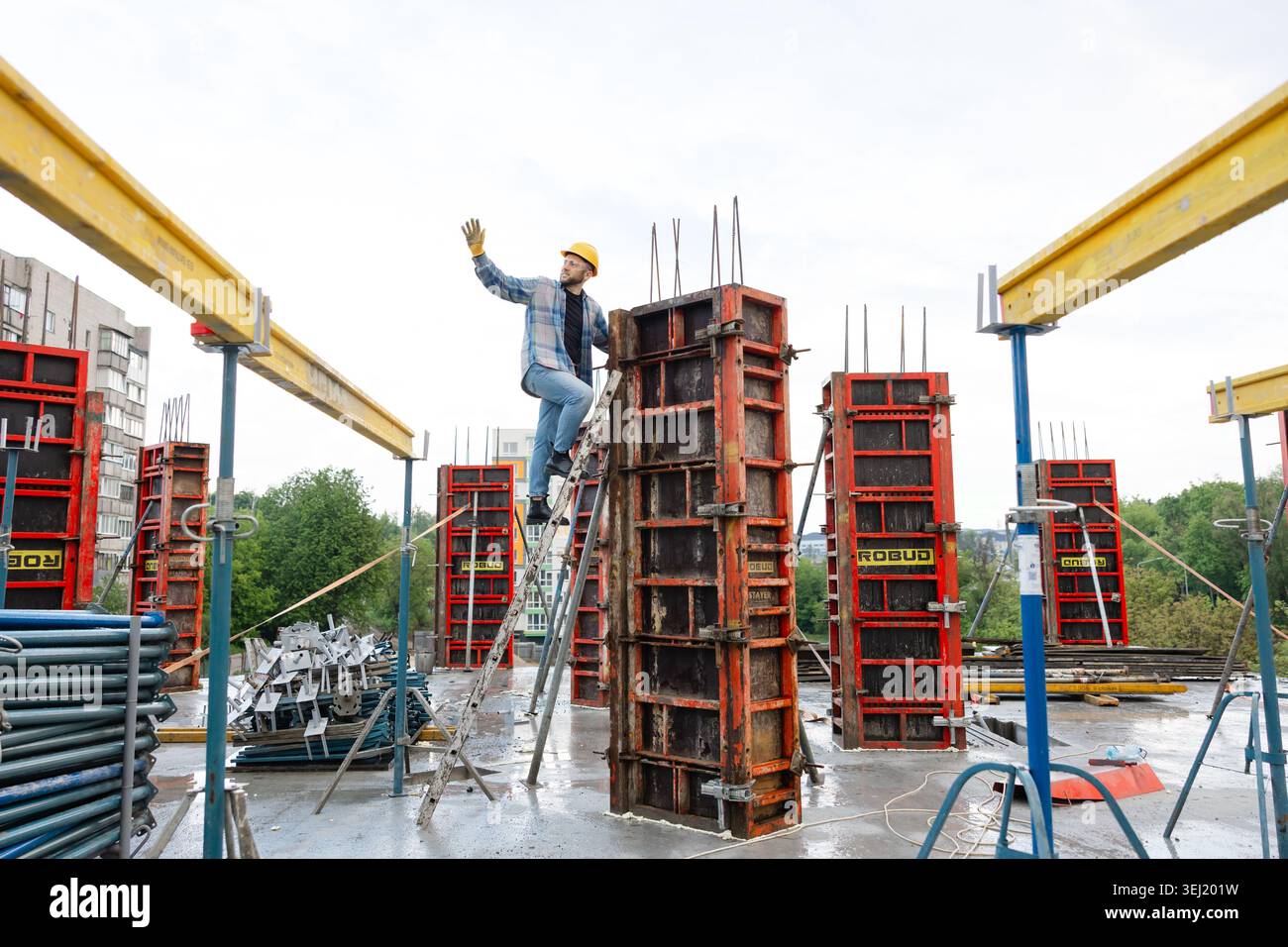 Construction worker in hard hat and safety glasses climbing ladder, setting metal formwork for a reinforced concrete column Stock Photo