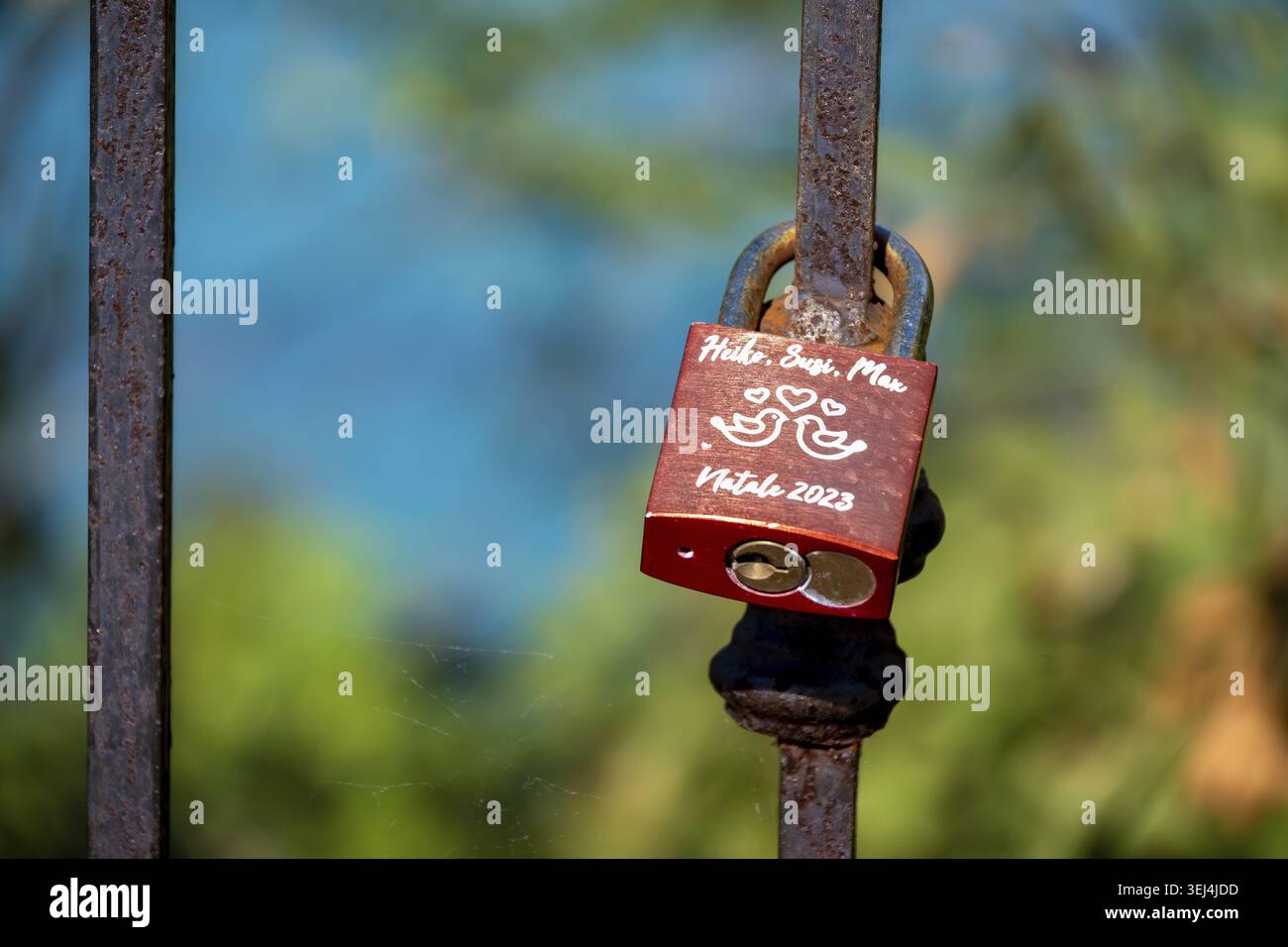 Vieste - Apulia - Italy - 08-29-2025: Red padlock with handwritten names and hearts hangs on a rusty iron fence, with blurred green and blue backgroun Stock Photo