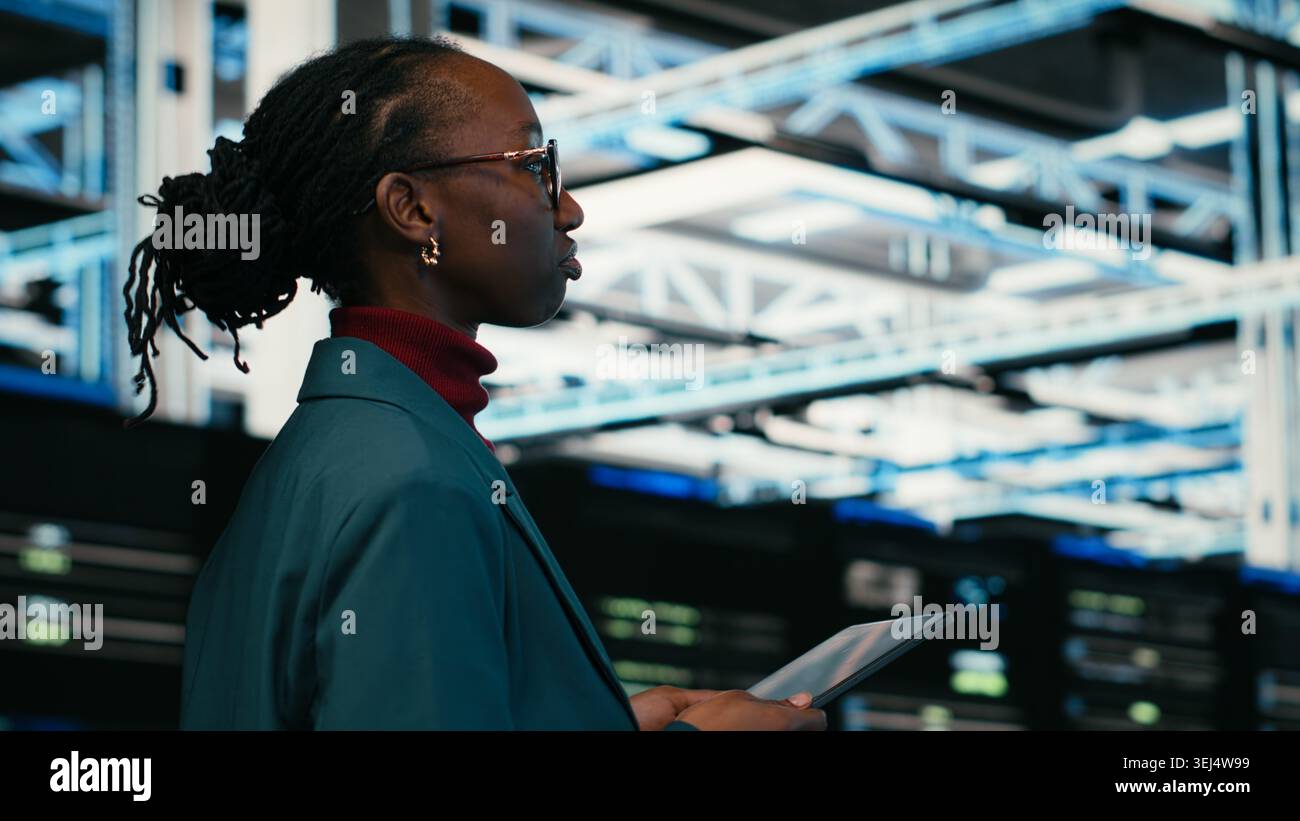 Data center programmer writing code for resource optimization using tablet application. African american woman walking in server hub using programming script on device to upgrade equipment, camera B Stock Photo