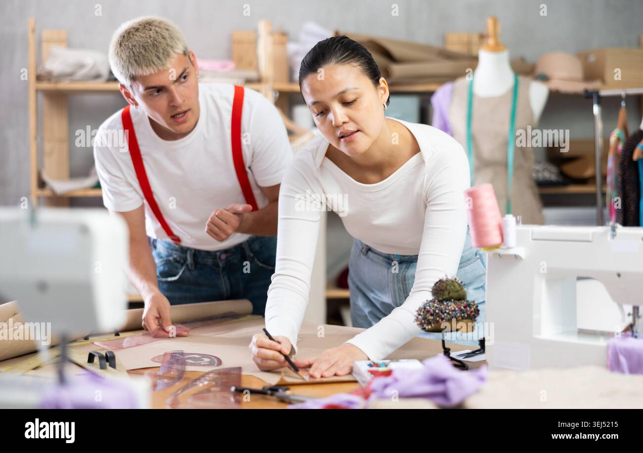 Two seamstresses male and female working in sewing workshop, woman drawing pattern on sheet of paper using curves and man consulting and helping Stock Photo