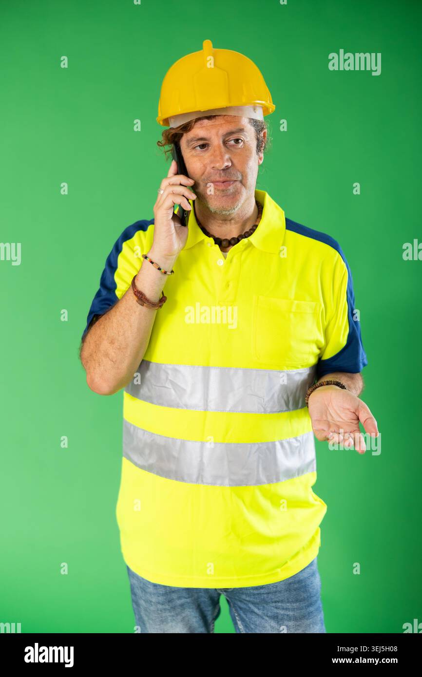 Adult man in reflective polo shirt and hard hat talking on mobile phone on green screen Stock Photo