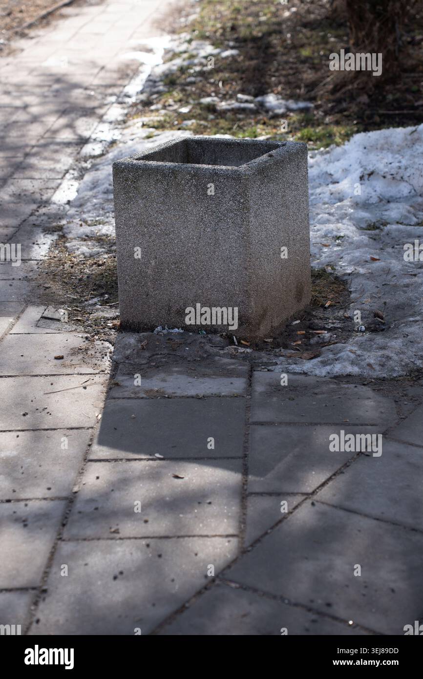 empty square concrete planter sitting on a gray urban sidewalk beside melting snow and scattered litter, casting angular shadows across paving stones; Stock Photo