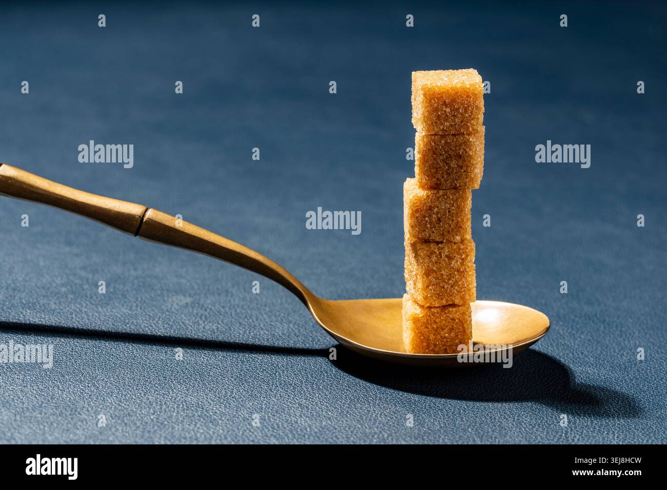 Tower of raw brown sugar cubes in a golden spoon over a blue textured surface, close up. Minimalist concept for sweetness portion control and diet Stock Photo