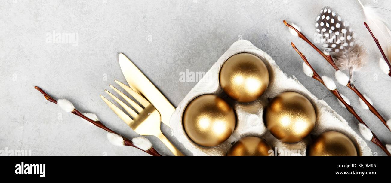Elegant Easter flat lay with golden eggs in carton, willow branches, feathers, bunny figurine and gold cutlery on light stone background. Festive spri Stock Photo
