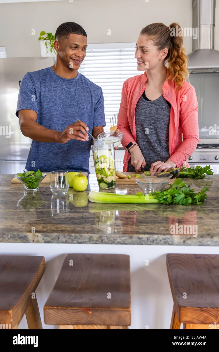Diverse couple in blue shirt, jacket using blender and chopping greens on granite island in kitchen Stock Photo