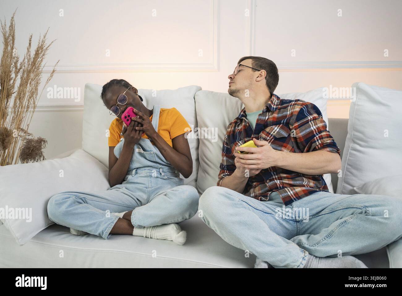 Multiethnic couple sitting on a white sofa, ignoring each other while using their smartphones, highlighting the impact of technology on modern relatio Stock Photo