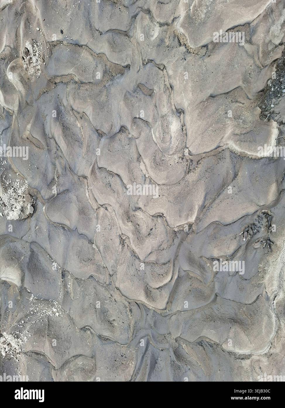 A top down view of natural grey sand ripples created by water flow at a river bank. The textures highlight the movement of water over time, displaying Stock Photo