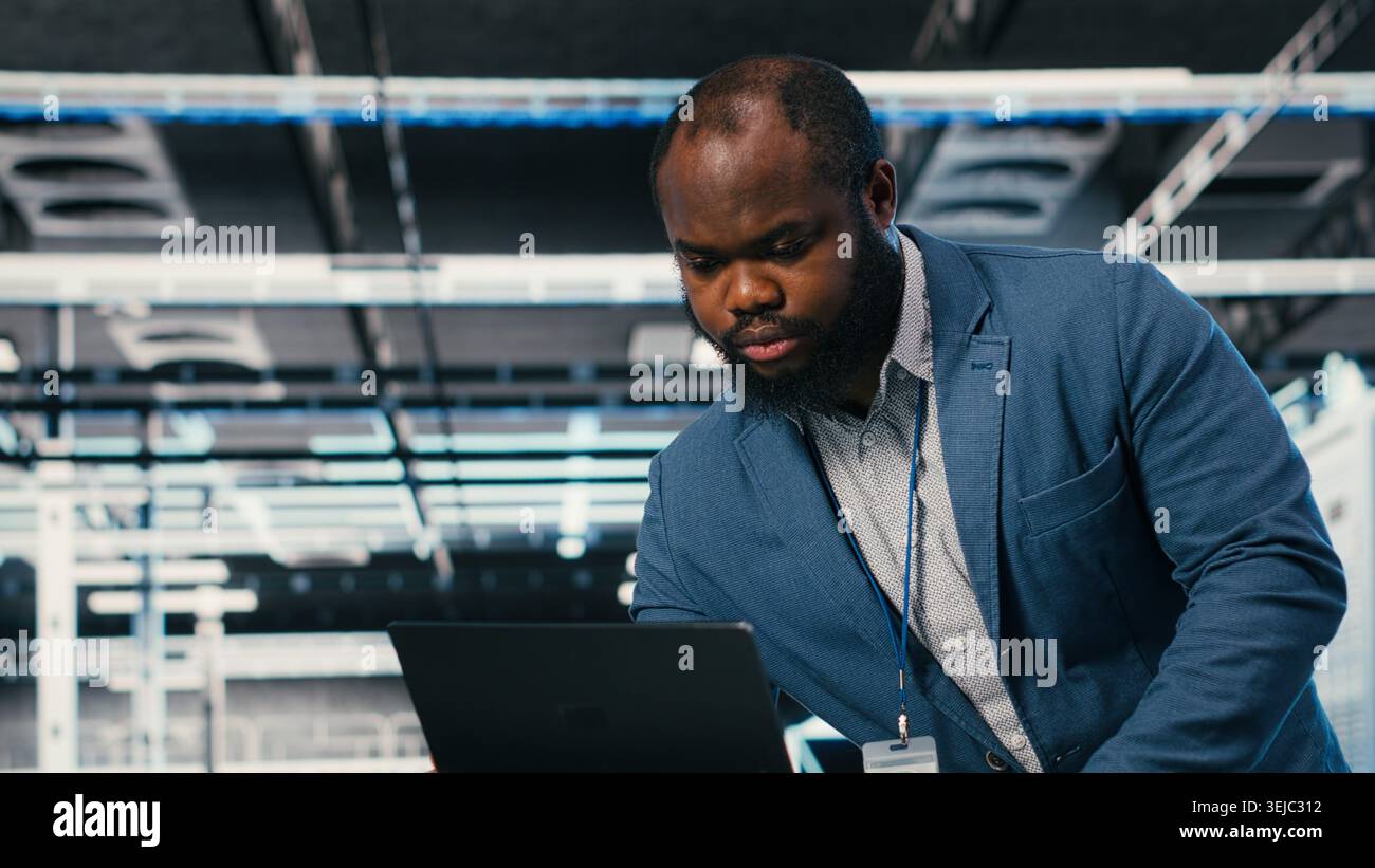 Programmer at server farm workplace desk using laptop, doing network management. Data center IT expert overseeing IT infrastructure supercomputers with software, typing on notebook keyboard, camera B Stock Photo