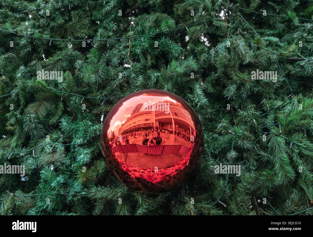 Large red Christmas ornament hangs on an artificial pine tree and reflects the Suria KLCC mall plaza and visitors in Kuala Lumpur, Malaysia Stock Photo