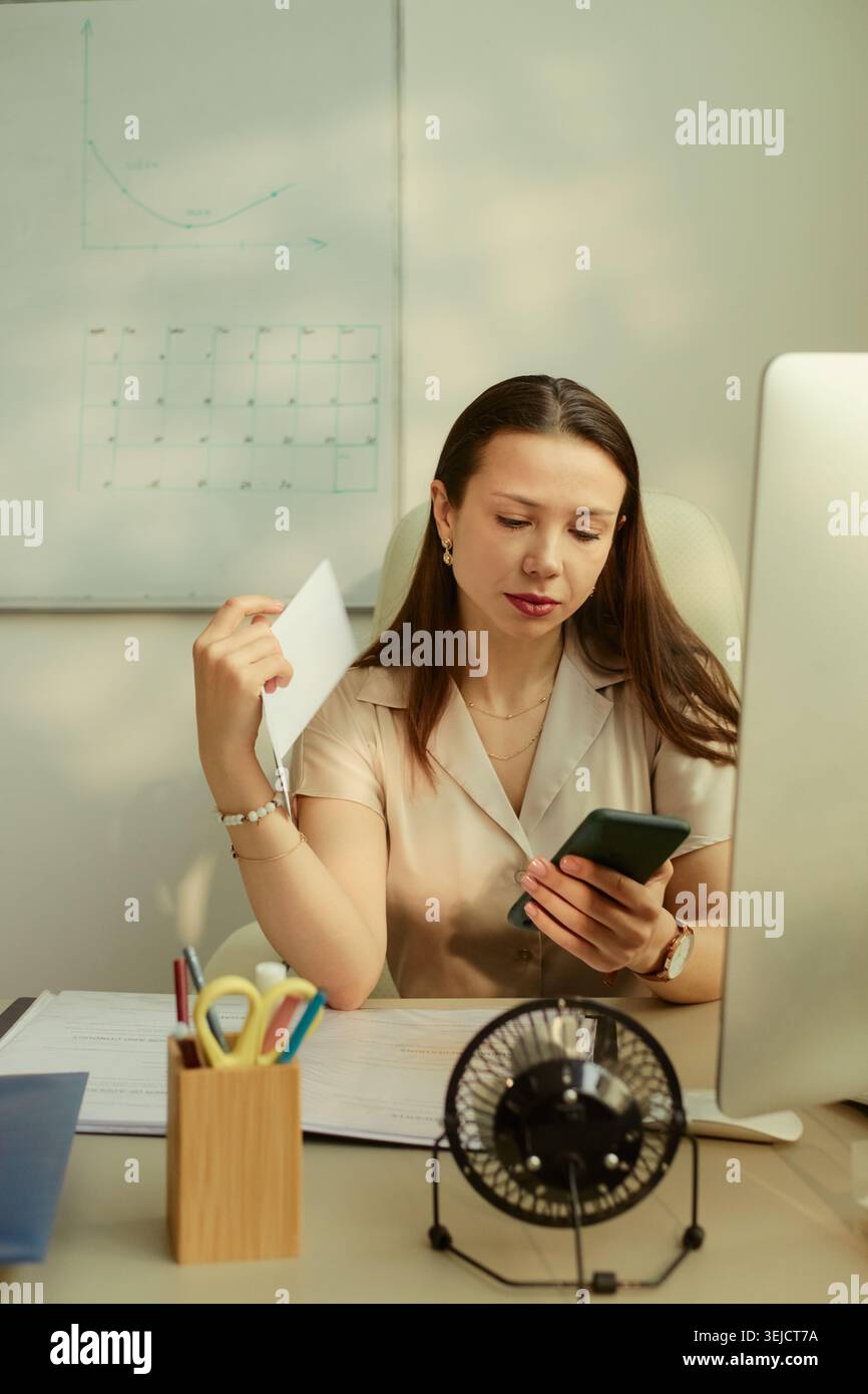 Young adult Caucasian businesswoman checking smartphone while holding document at desk, managing office workflow and communication. Useful for business productivity or admin content Stock Photo