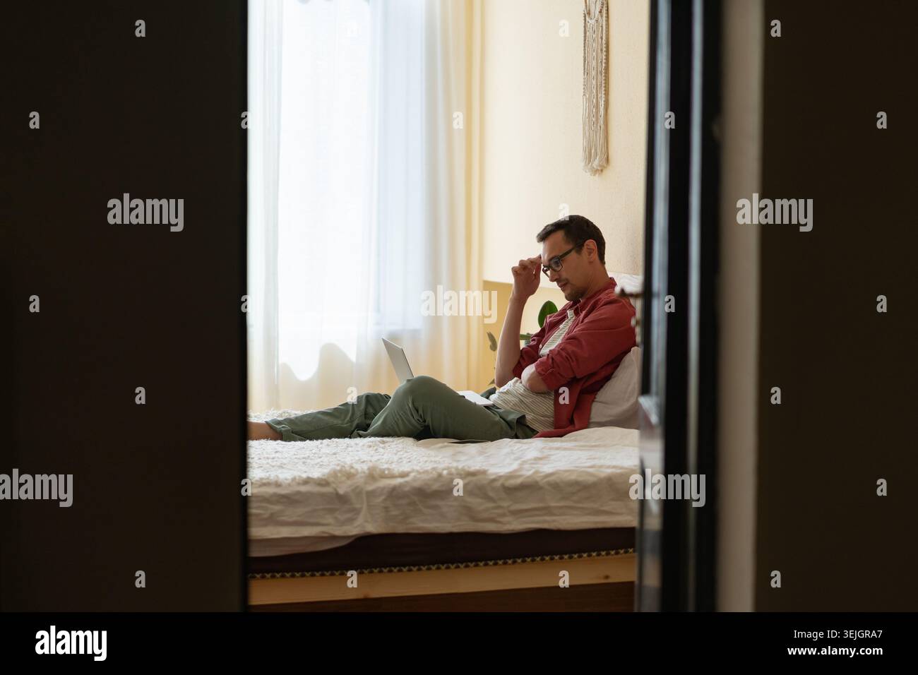 Thoughtful man freelancer sitting on bed with laptop, stuck with task while working remotely Stock Photo