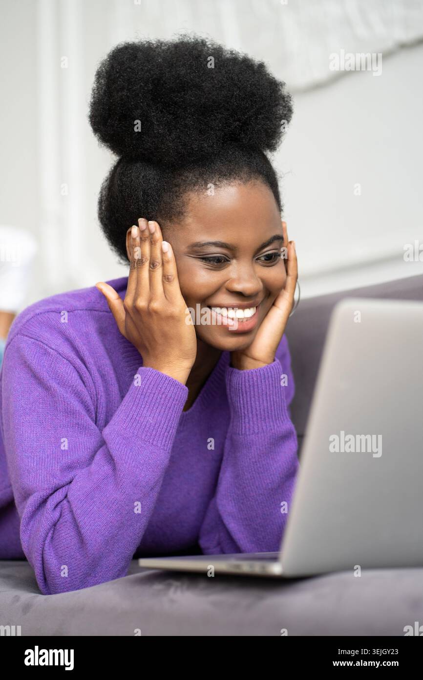 Smiling Afro-American millennial woman in purple sweater lying on couch, looking at camera webcam and talking on a video call or Stock Photo