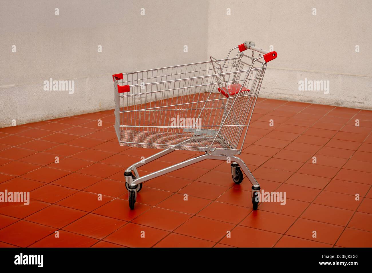 An empty steel supermarket trolley standing alone on an orange tiled floor against a white wall Stock Photo