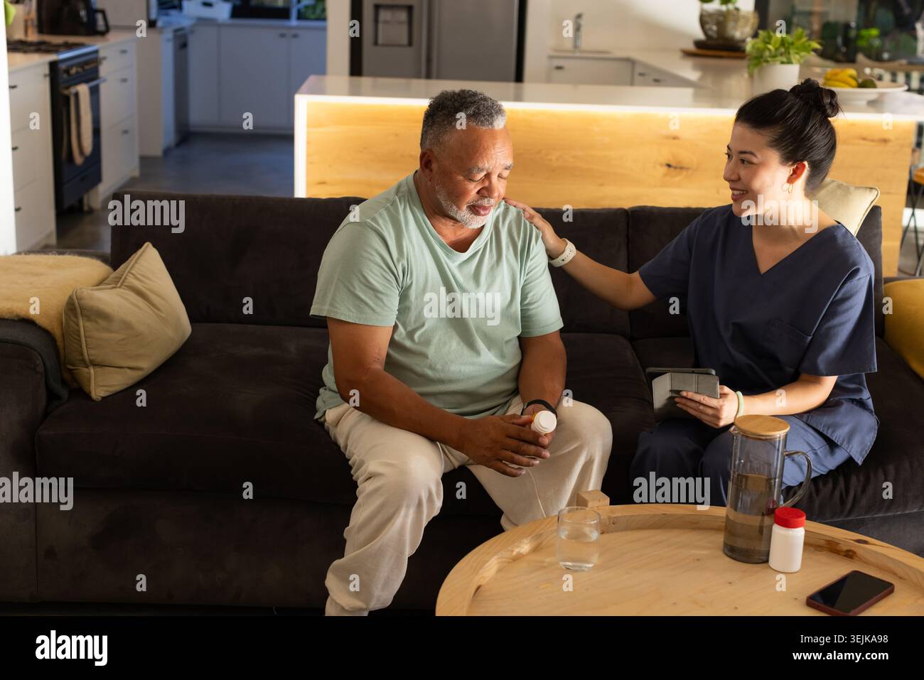 African American senior holding pill bottle while Asian caregiver navy scrubs using tablet on sofa Stock Photo