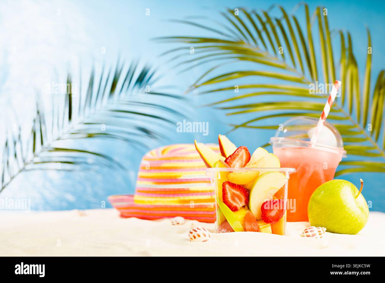 Resort lifestyle - picnic with cold cocktail, fresh tropical fruit salad, sun hat on white sand with palm, sun glare in blue sea Stock Photo