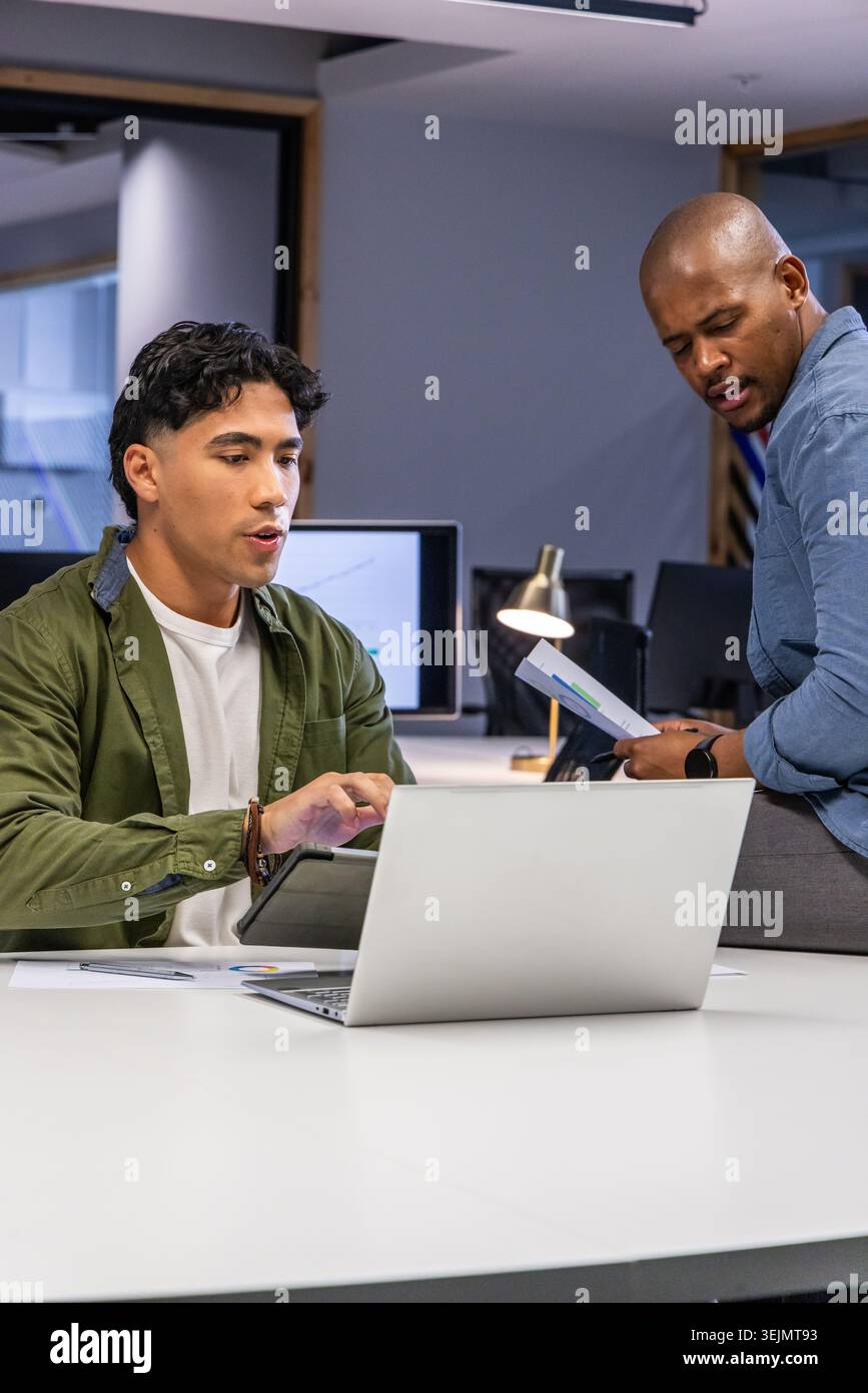 Diverse coworkers collaborating, reviewing printed charts and tablet using laptop at meeting table. Teamwork, graphs, reports, o Stock Photo